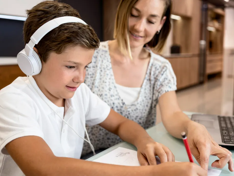 A parent sits beside her son as he works at a laptop wearing headphones
