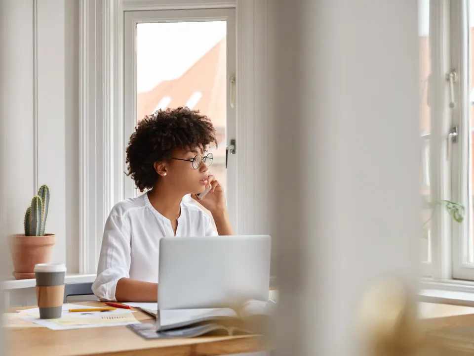 A tutor on a phone call at a desk with a laptop and notes by a bright window